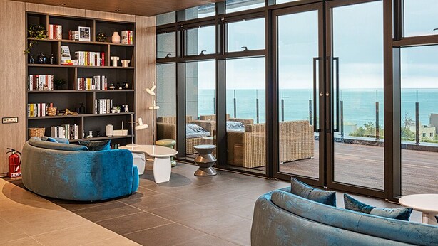 Una sala de estar moderna con una amplia estantería, asientos cómodos y vista al mar a través de grandes ventanales.