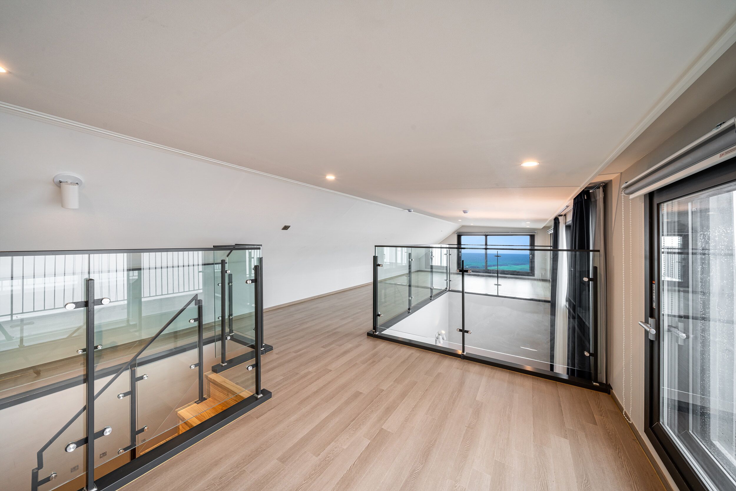 Un interior espacioso con piso de madera, barandillas de vidrio y vista al mar a través de amplios ventanales.