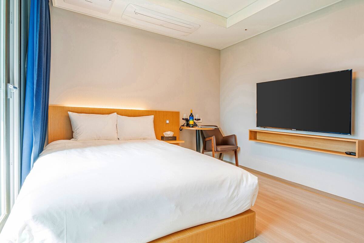 Habitación de hotel con una cama grande, un escritorio con silla, un televisor de pantalla plana en la pared y una ventana con cortinas.
