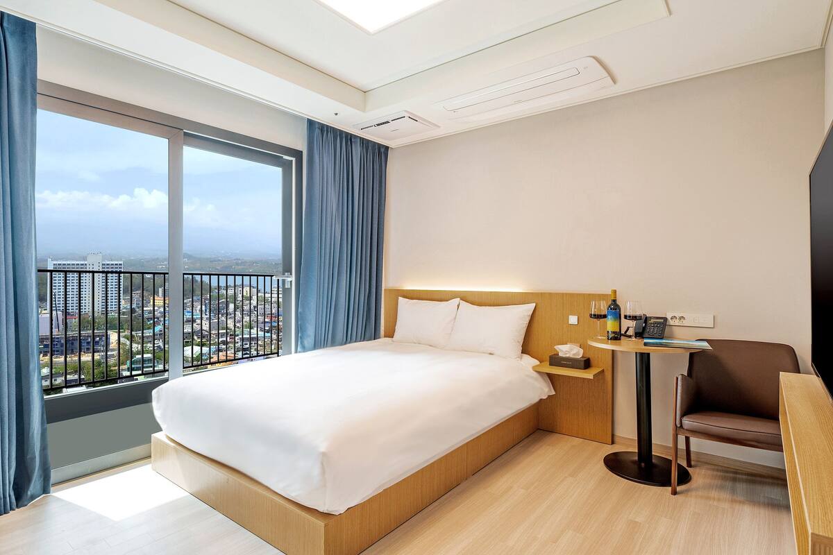 Habitación de hotel con una cama grande, una mesita con una silla, vistas de la ciudad a través de un ventanal amplio y un balcón.