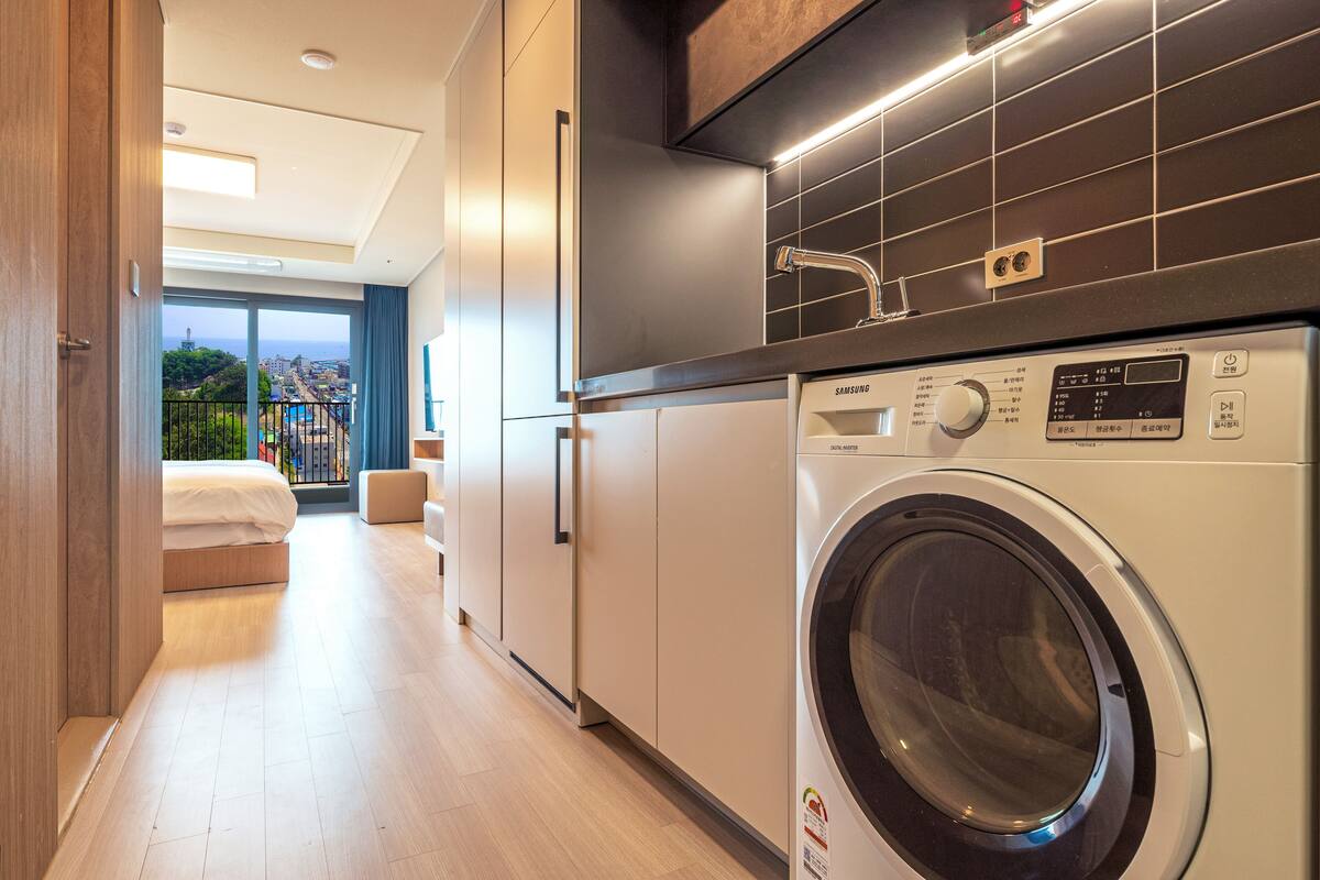 Una cocina moderna con lavadora y secadora integradas, un amplio ventanal con vistas a la ciudad y un pasillo espacioso que conduce a un dormitorio.