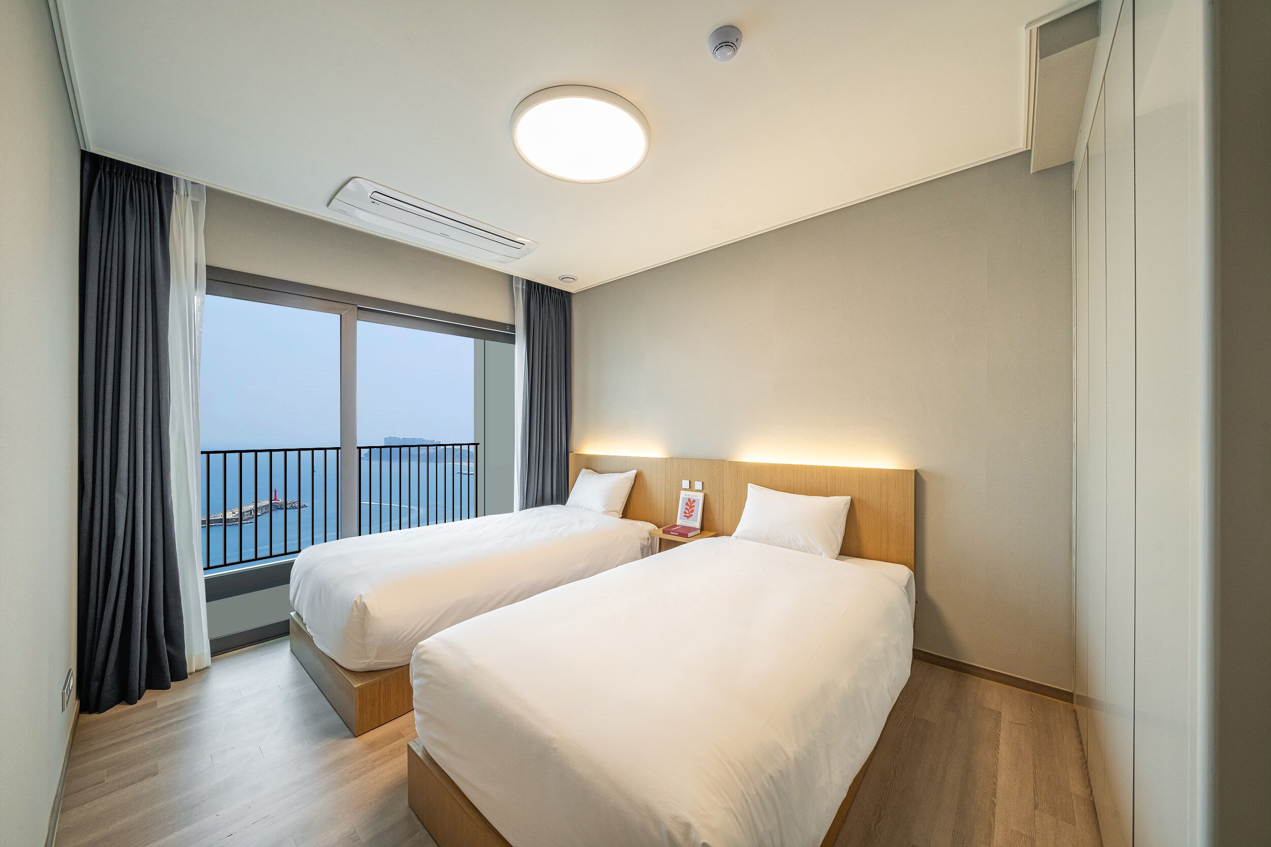 A hotel room with two beds, a large window with a view of the sea, and a ceiling light.