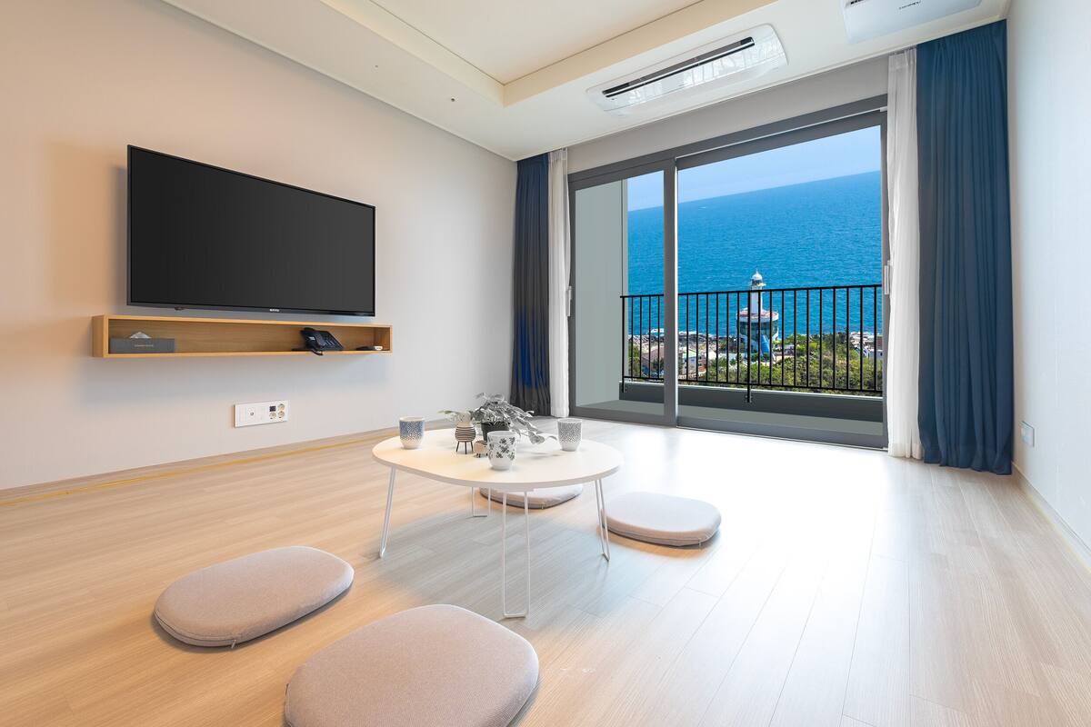 Una habitación de hotel moderna con un televisor grande, un balcón con vistas al mar, una mesa redonda con sillas y un pequeño sofá.