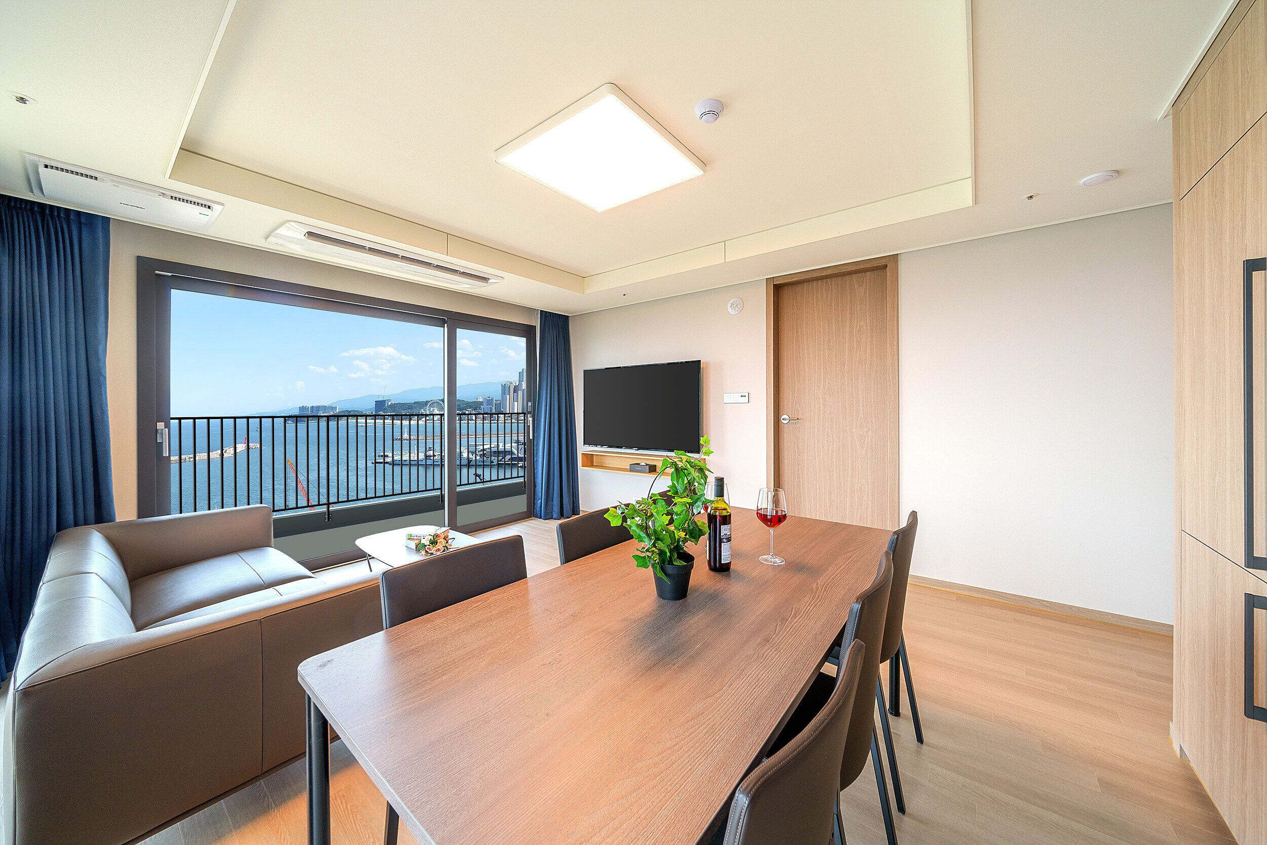 A modern hotel room with a large window, a balcony, a wooden dining table, and a flat-screen TV.