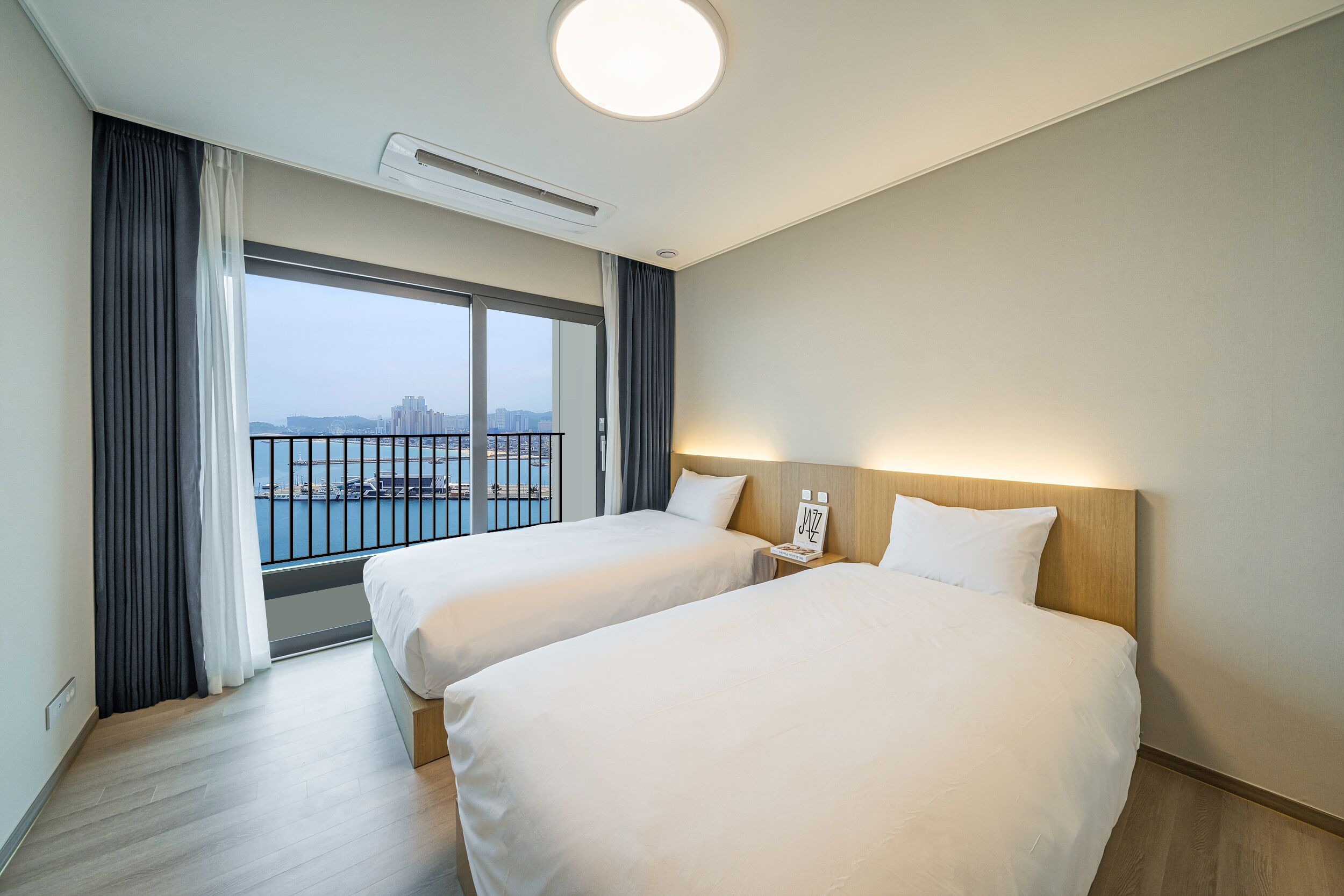 A hotel room with two beds, a large window with a view of the city, and a ceiling light.