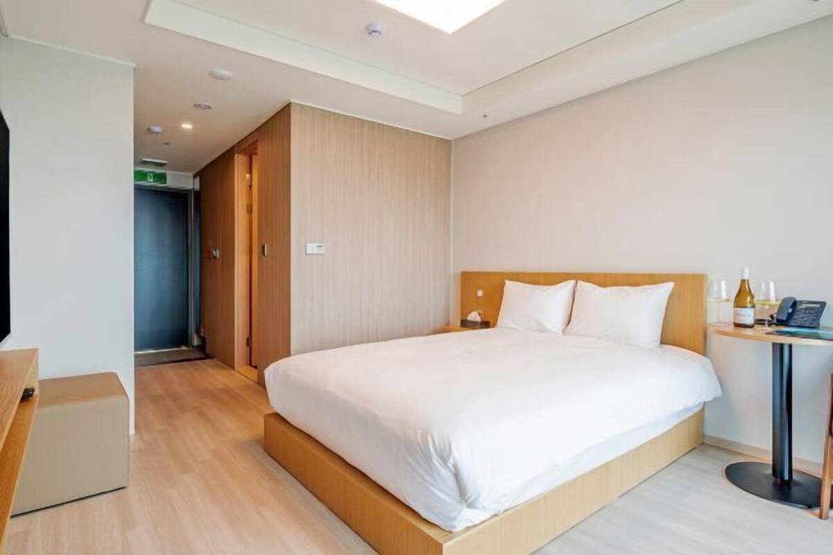Una habitación de hotel con una cama grande, un cabecero de madera, una mesita con un teléfono y una botella, y un armario.