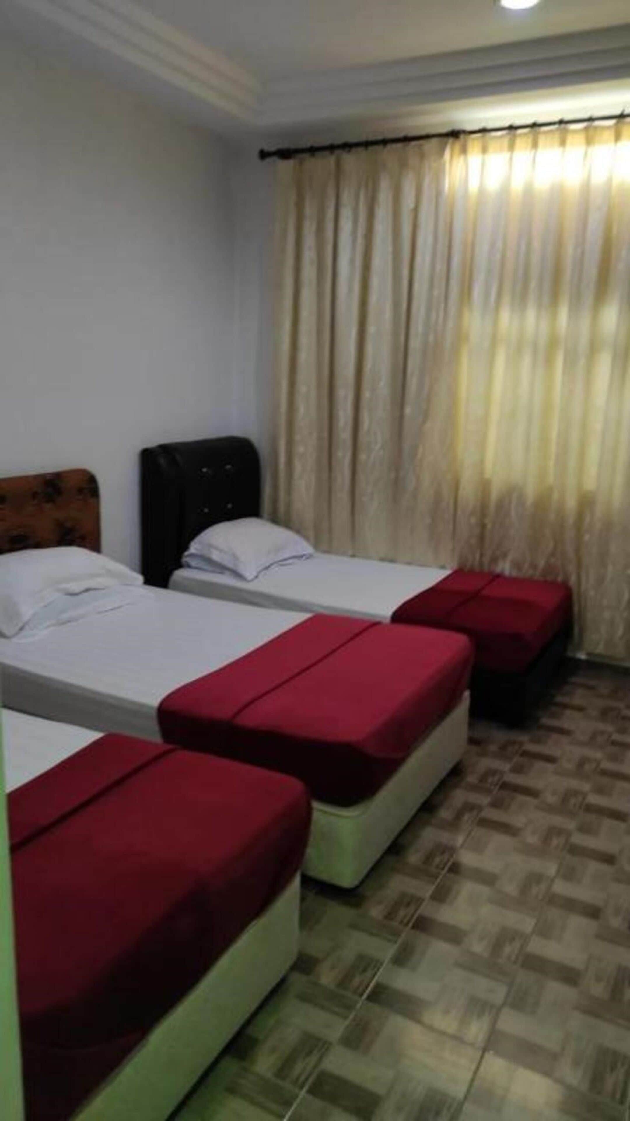 A hotel room with two beds, each with a red bedspread, and a large window with beige curtains.