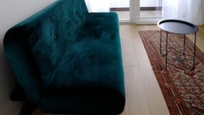 Un sofá azul verdoso en una habitación con suelo de madera, una mesita redonda pequeña y una alfombra estampada.