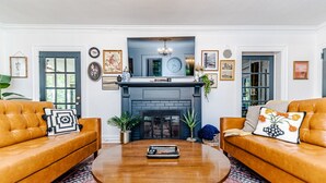 Sala de estar com dois sofás de couro, uma mesa de centro redonda de madeira, uma lareira e diversas molduras com imagens nas paredes.