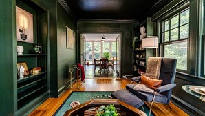 Uma sala de estar aconchegante com parede verde escura, piso de madeira e um espaço de estar confortável, com uma poltrona e um sofá. Uma mesa de jantar com cadeiras aparece ao fundo, e uma estante está repleta de diversos objetos.