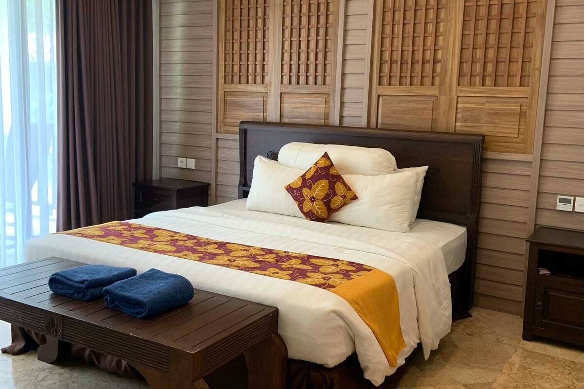 Ein Hotelzimmer mit einem großen Bett, einem Nachttisch aus Holz, einem Tisch mit Handtüchern und einem Fenster mit Vorhängen.