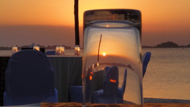 A sunset view with a glass table in the foreground, a wooden post, and chairs with blue covers. - lorin beach resort belitung (Tanjung Pandan)