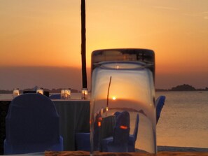 Zonsondergang met een glazen tafel op de voorgrond, een houten paal en stoelen met blauwe bekleding.