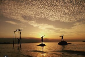 Ein Sonnenuntergang über einem ruhigen Meer, mit zwei Personen auf kleinen Inseln: eine auf einer Schaukel, die andere auf einem Felsen.