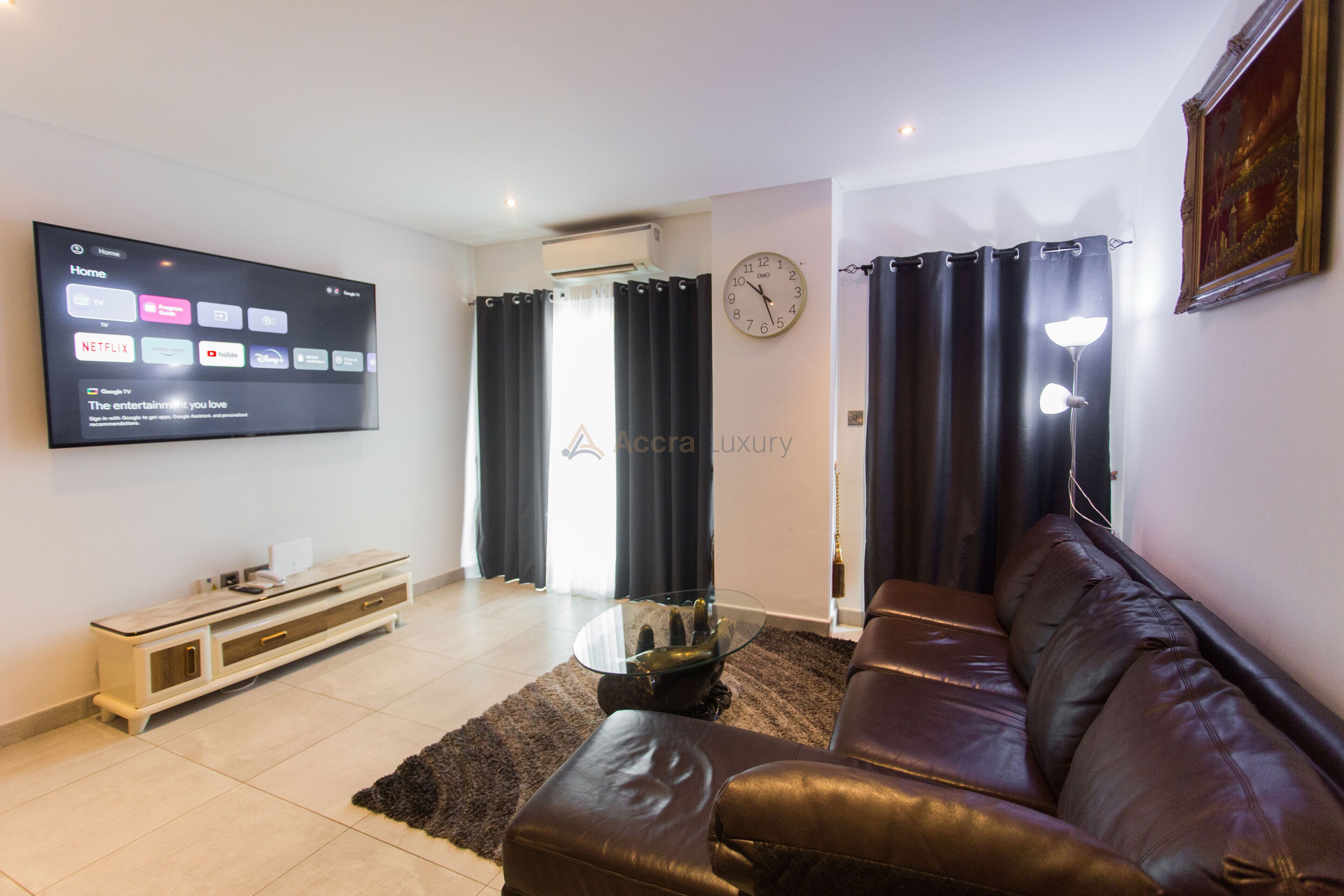 A modern living room with a leather sofa, a glass coffee table, a flat-screen TV displaying streaming services, and a wall clock.