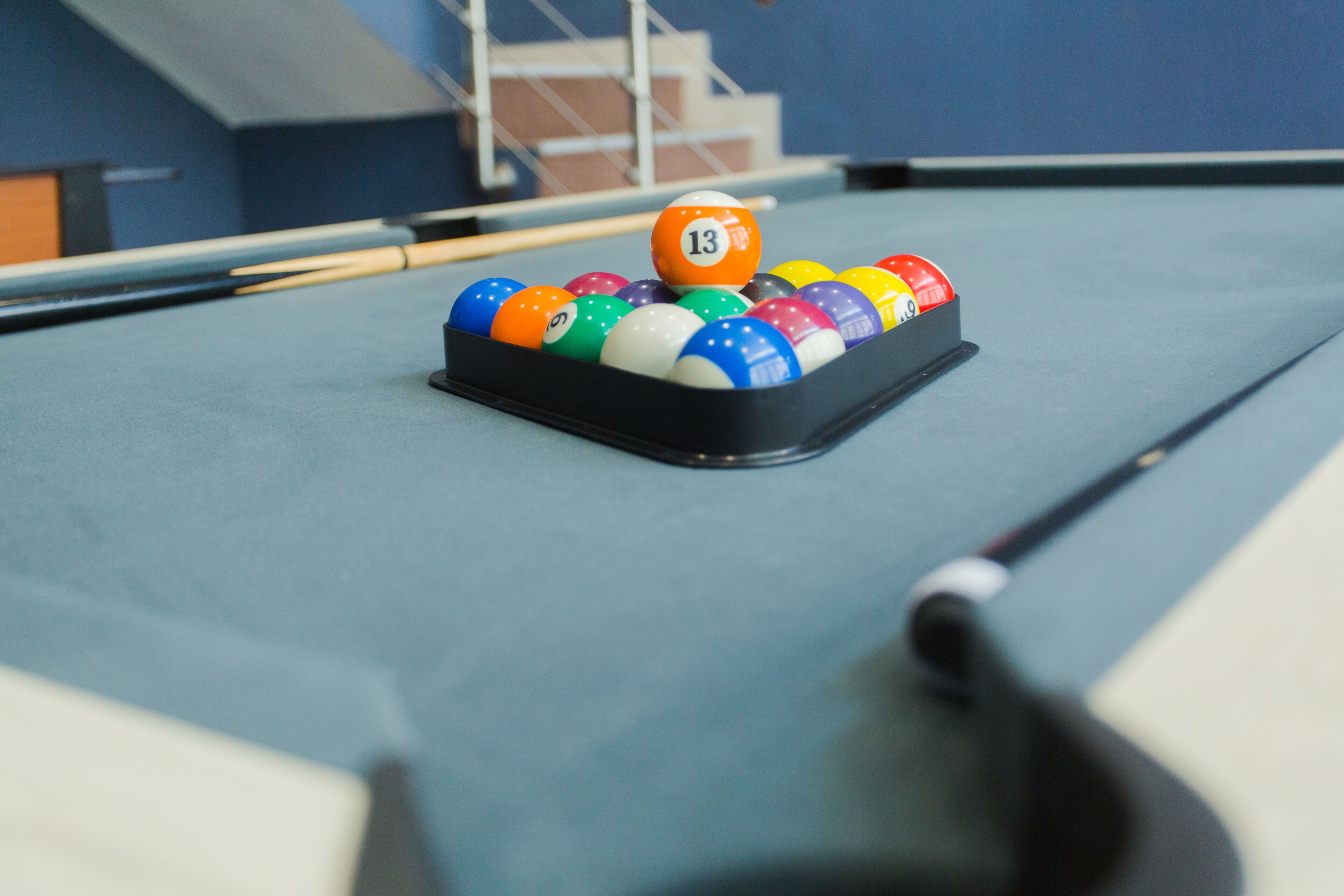 Una mesa de billar con un repuesto de bolas de billar, incluyendo la bola número 13, en una habitación con paredes azules y una escalera al fondo.