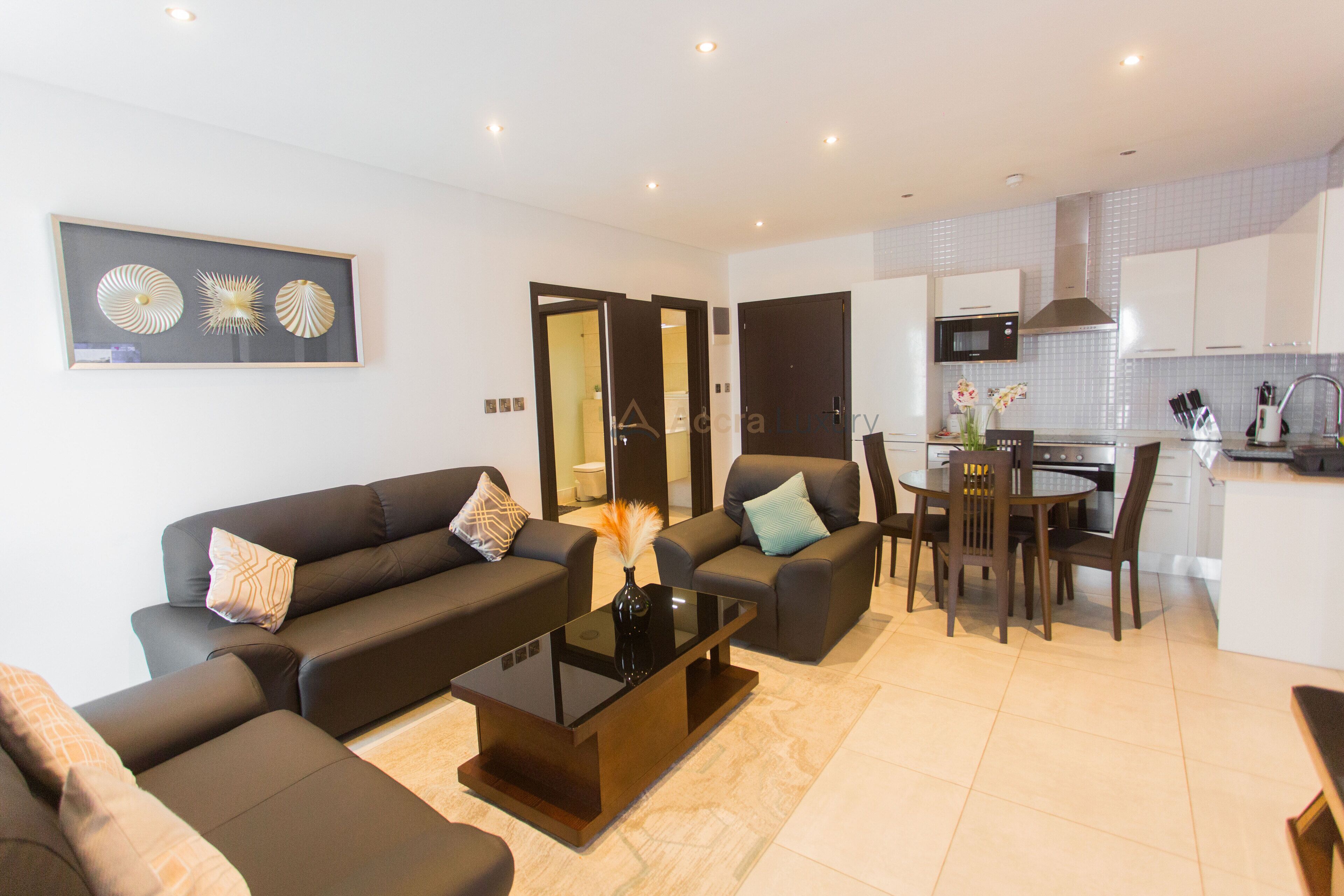 A modern living room with a sofa set, a coffee table, and a dining area with a table and chairs. There is a kitchen area with cabinets and a microwave.