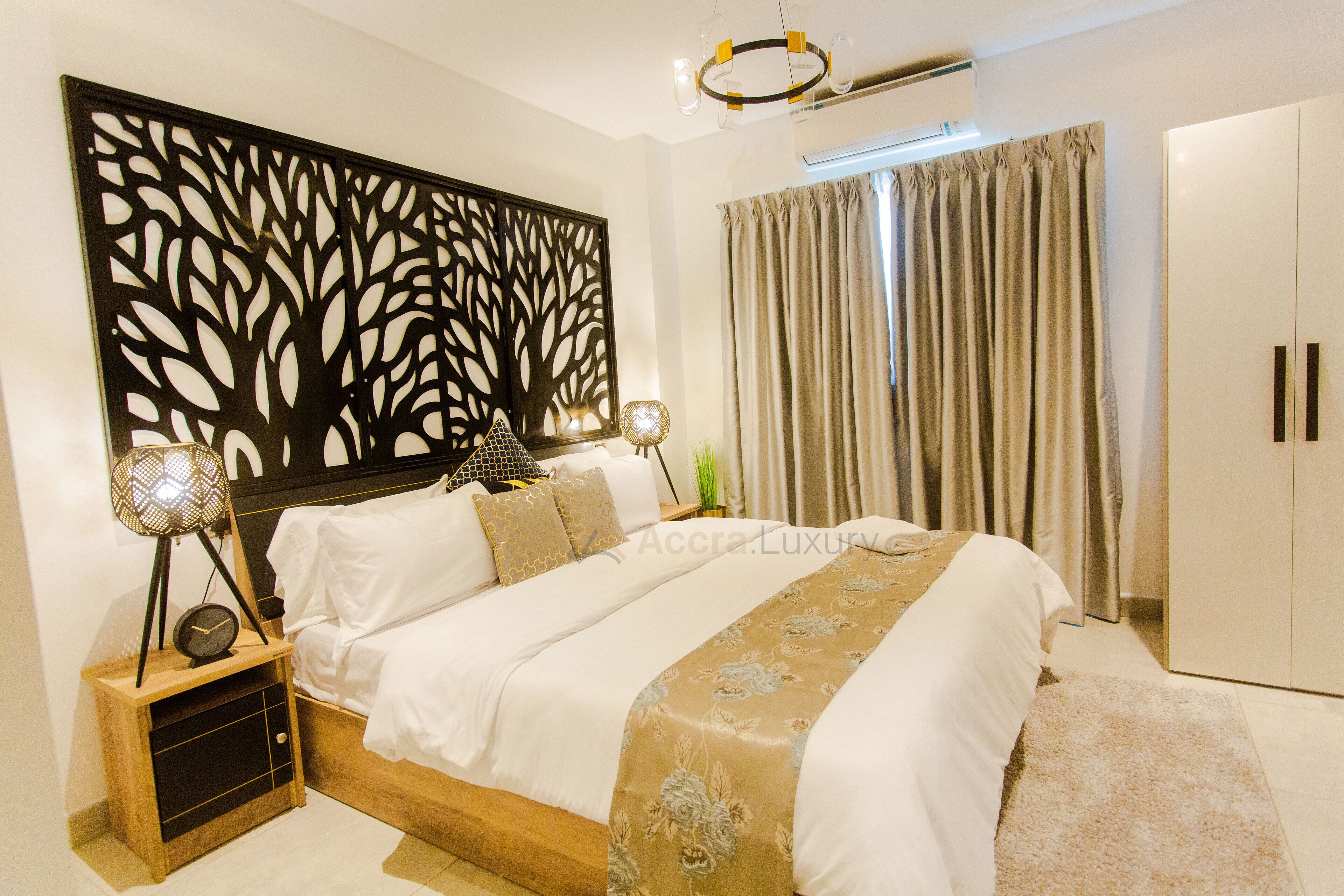 A modern bedroom with a large bed, a decorative wall panel, a wardrobe, and a bedside table with a lamp.