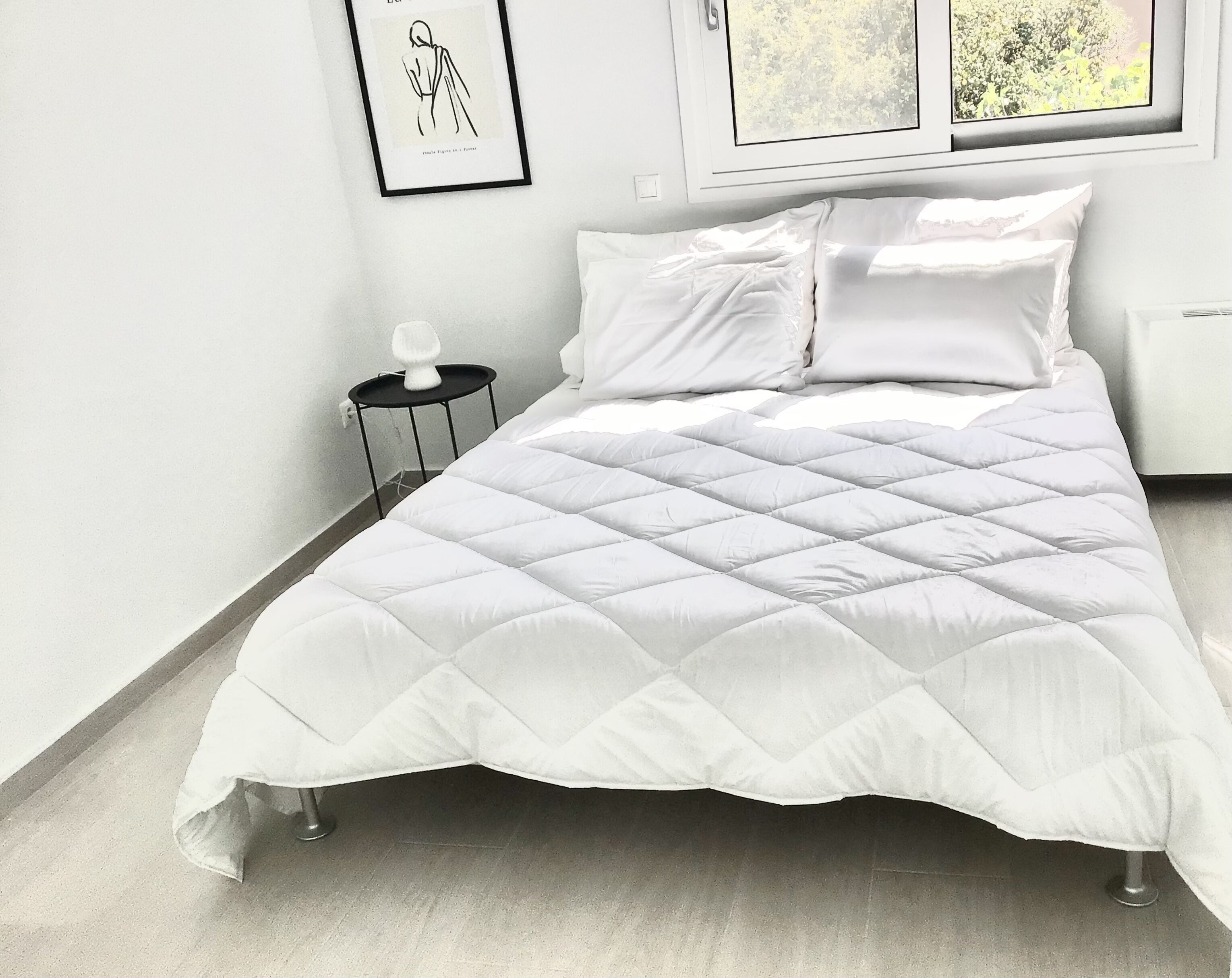 Un lit bien fait, avec une couette blanche matelassée, une petite table de chevet surmontée d’une lampe blanche et un tableau encadré accroché au mur.