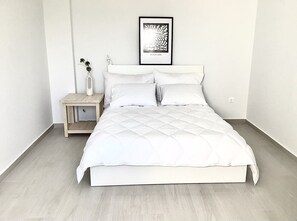 A neatly made bed with white bedding in a room with light wooden flooring and white walls.
