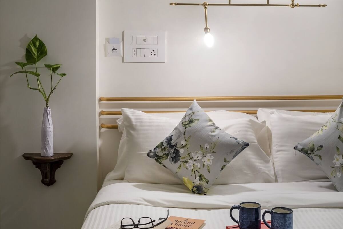 A neatly made bed with pillows, a bedside table with a plant, a book, and two mugs on a tray.