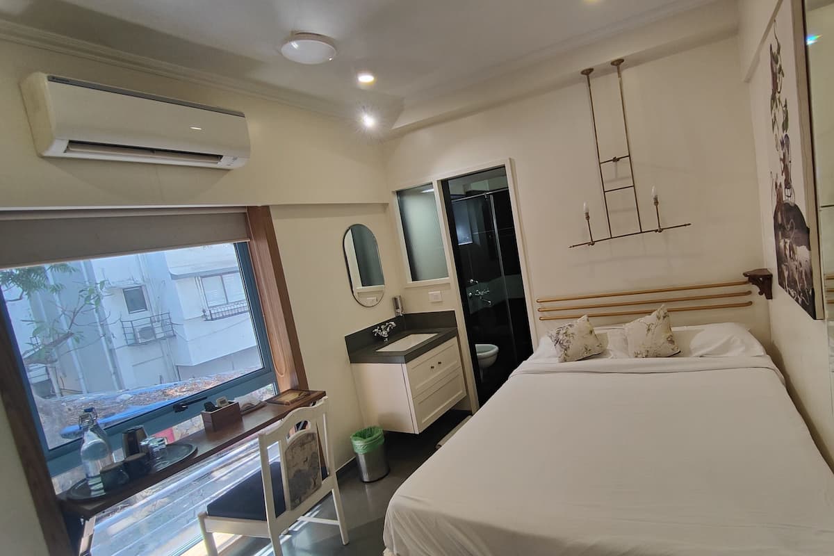 A bedroom with a bed, a desk with a chair, a window, an air conditioning unit, and a bathroom with a toilet and sink.
