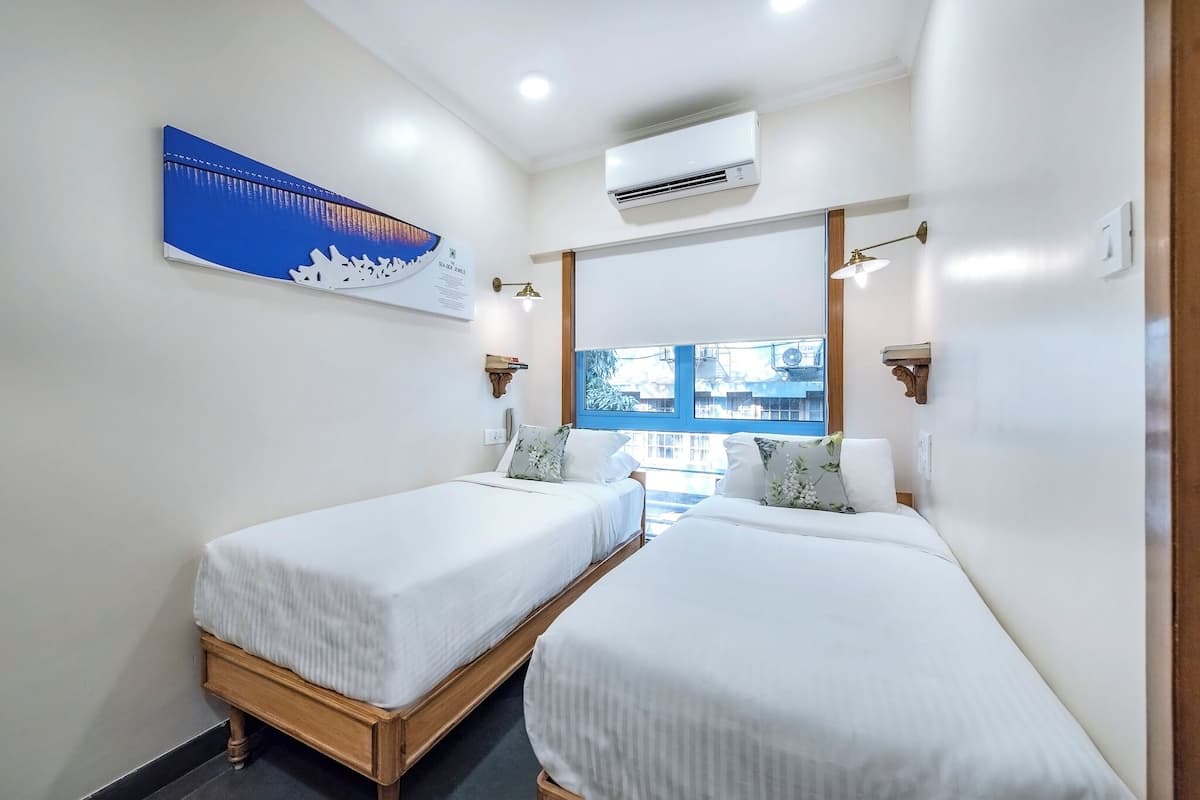 A hotel room with two single beds, a large window, an air conditioning unit, and a wall-mounted artwork.