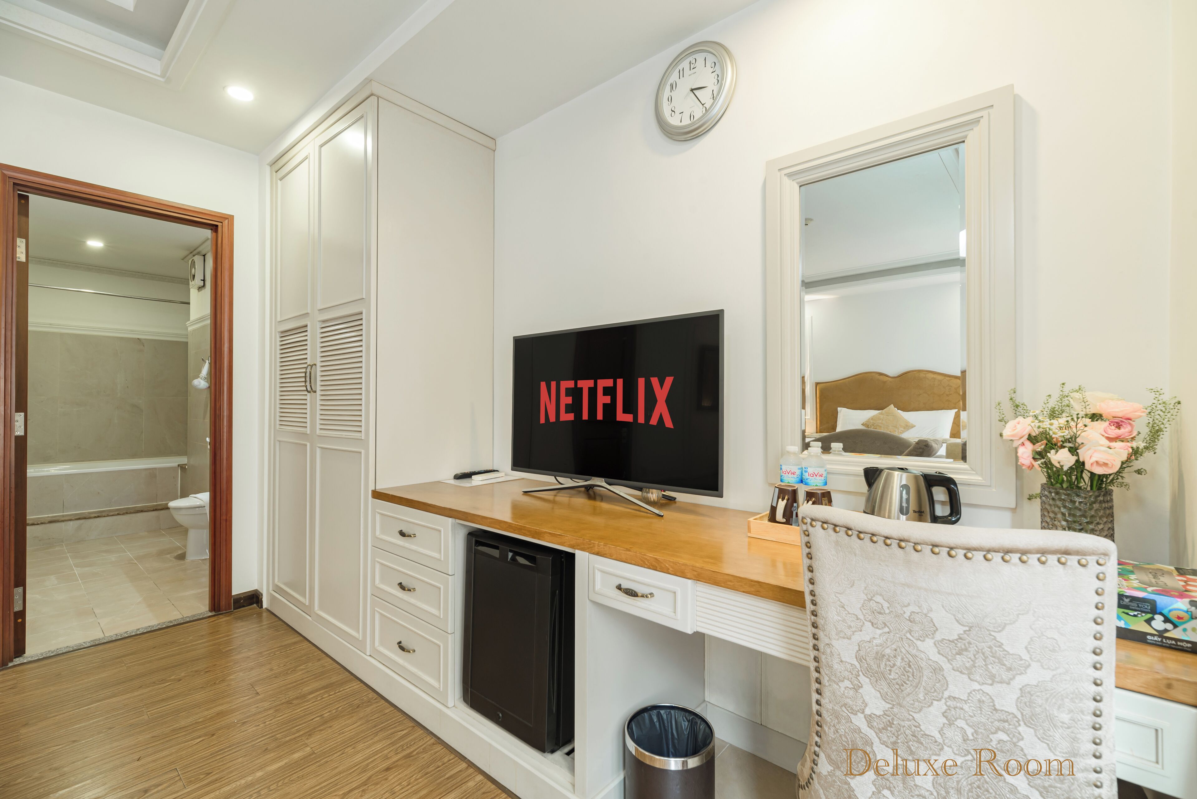 A room with a wooden desk, a television displaying 'NETFLIX', a clock, a mirror, and a trash bin.