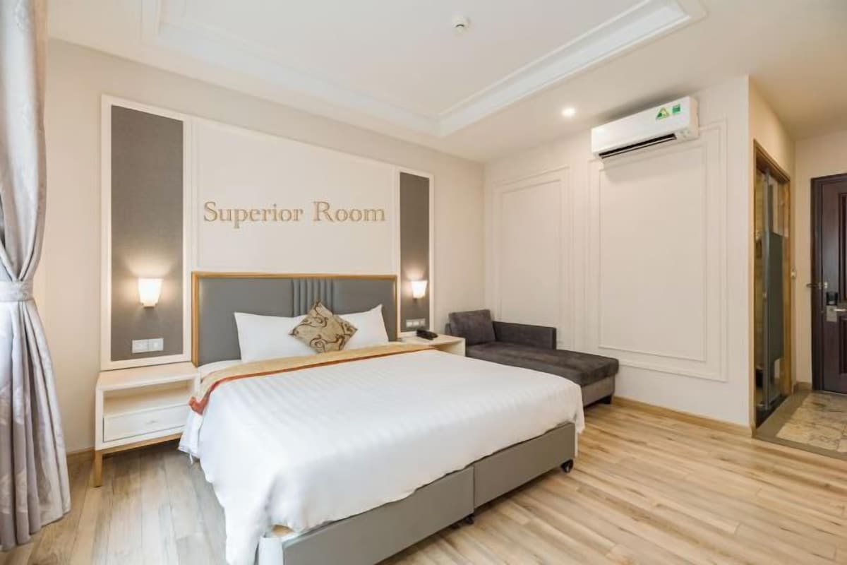 Una habitación de hotel con cama, mesitas de noche, un sofá gris y un equipo de aire acondicionado.
