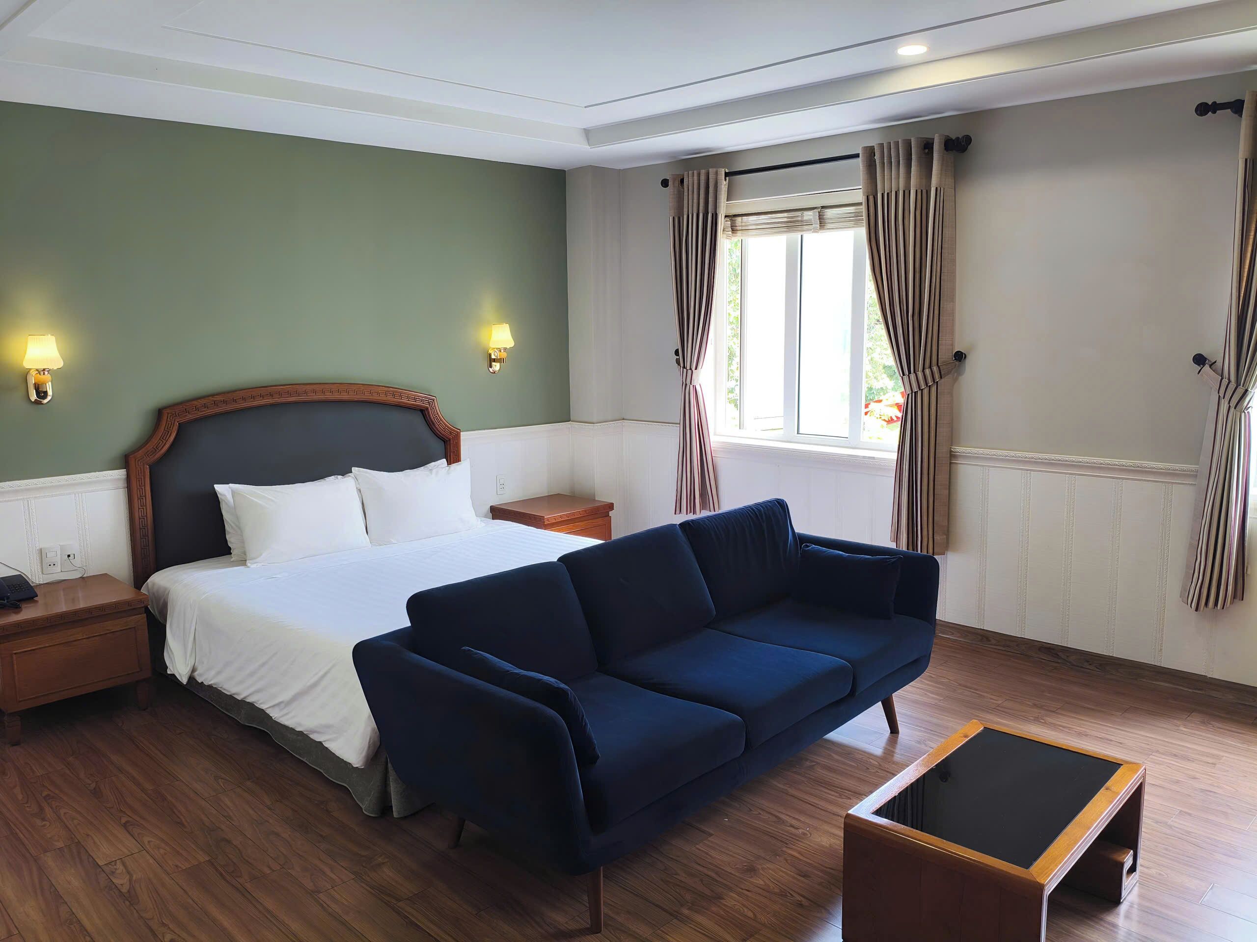 A hotel room with a bed, a blue sofa, a wooden nightstand, a small table, and a window with curtains.