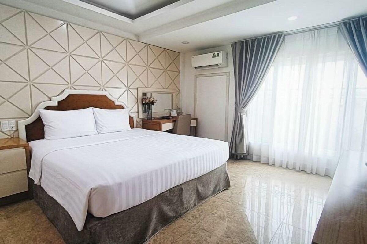 A hotel room with a large bed, a nightstand, a desk, and a window with curtains.