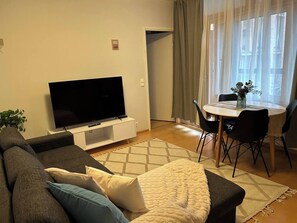 A living room with a flat-screen TV, a sofa, a dining table with chairs, and a window with curtains.