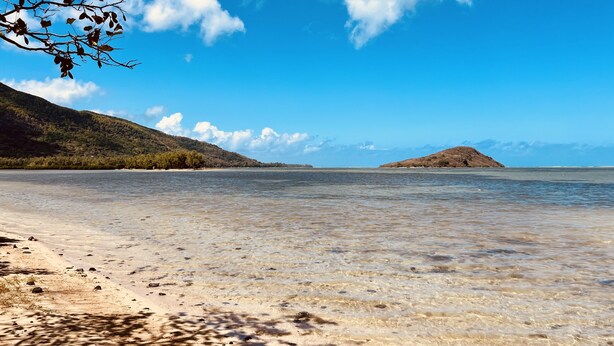 Una playa con aguas poco profundas y cristalinas, una orilla arenosa y colinas a lo lejos.