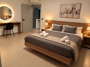 A hotel room with a bed, a desk, a chair, a bedside table with a lamp, and a wall-mounted painting.