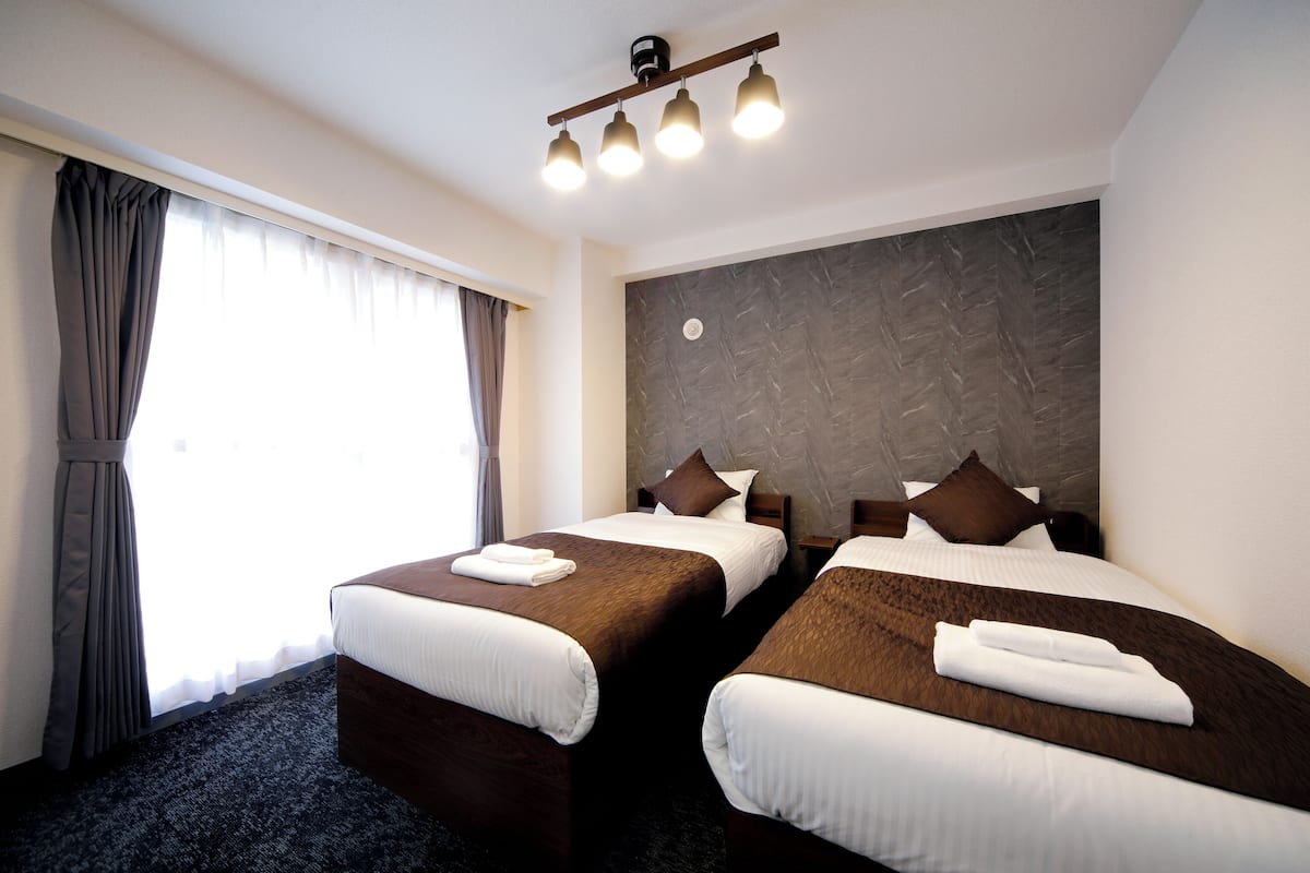 Habitación de hotel con dos camas, un ventanal grande con cortinas, un cabecero moderno y una lámpara de techo.