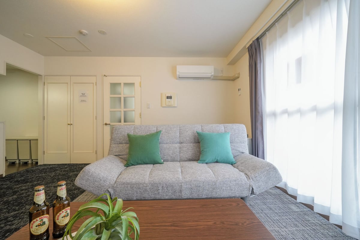 Una sala de estar con un sofá gris, dos cojines verdes, una mesa de centro de madera y una planta en maceta. Hay una ventana con cortinas, un equipo de aire acondicionado en la pared y una puerta con un panel de vidrio esmerilado.