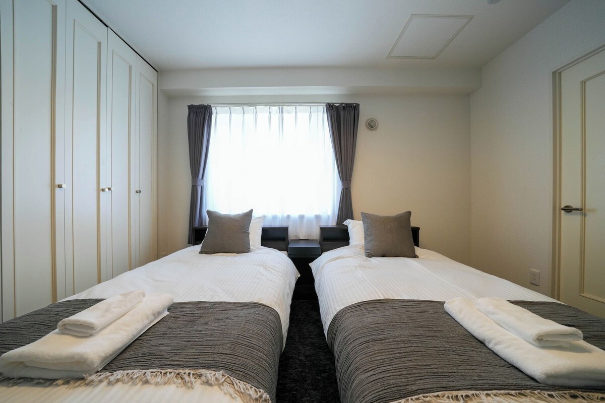 A hotel room with two single beds, a large window with curtains, a wardrobe, and a door.