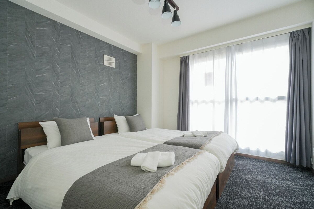 A hotel room with a large bed, a grey textured wall, and a window with grey curtains.