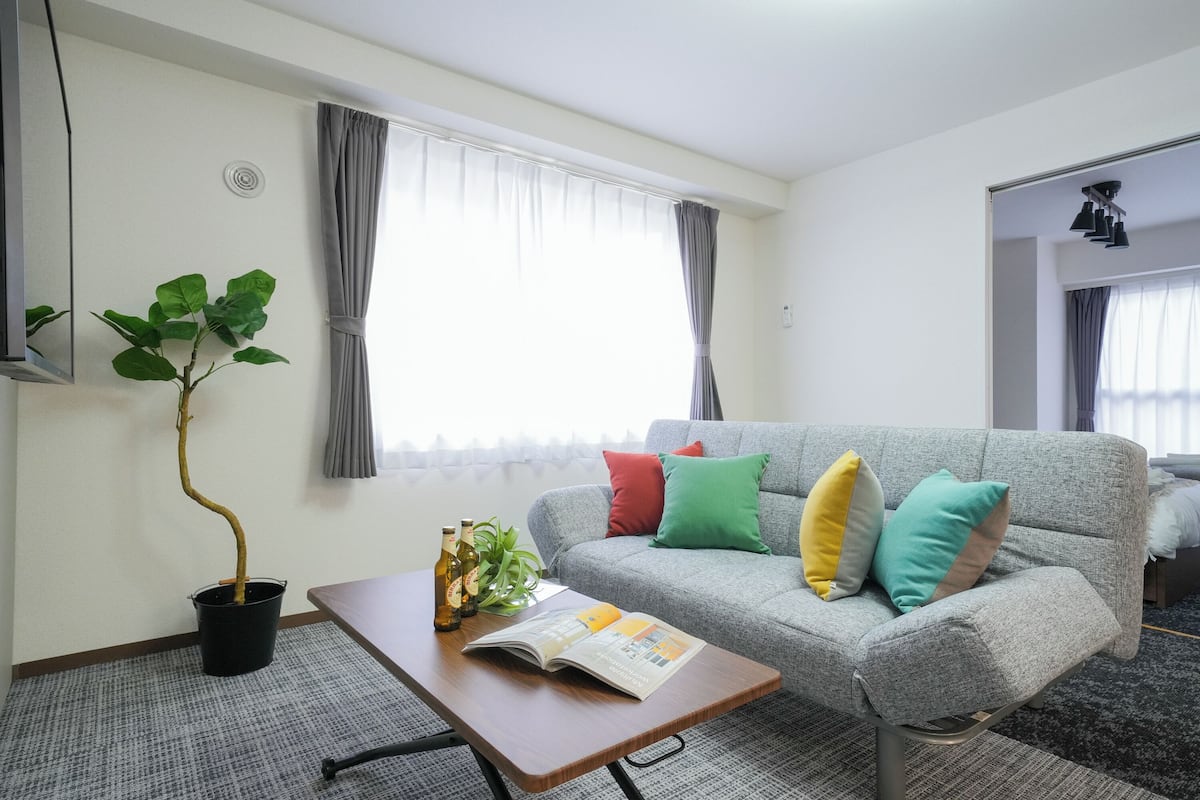 Habitación de hotel con un sofá gris, cojines de colores, una mesa de centro de madera con una revista, una planta en maceta y una ventana con cortinas.