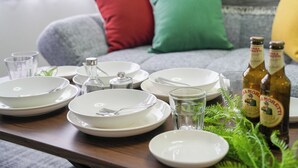 Ein Esstisch ist gedeckt mit weißen Tellern, Gabeln und Gläsern, dazu liegen zwei Bierflaschen bereit. Im Hintergrund sieht man eine Couch mit Kissen.