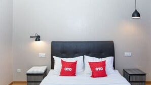 Un lit bien fait, avec une literie blanche et deux oreillers rouges sur lesquels est inscrit le mot « OYO ». Au-dessus du lit, on aperçoit une suspension noire, et à côté, une petite table de chevet surmontée d’un coussin blanc.