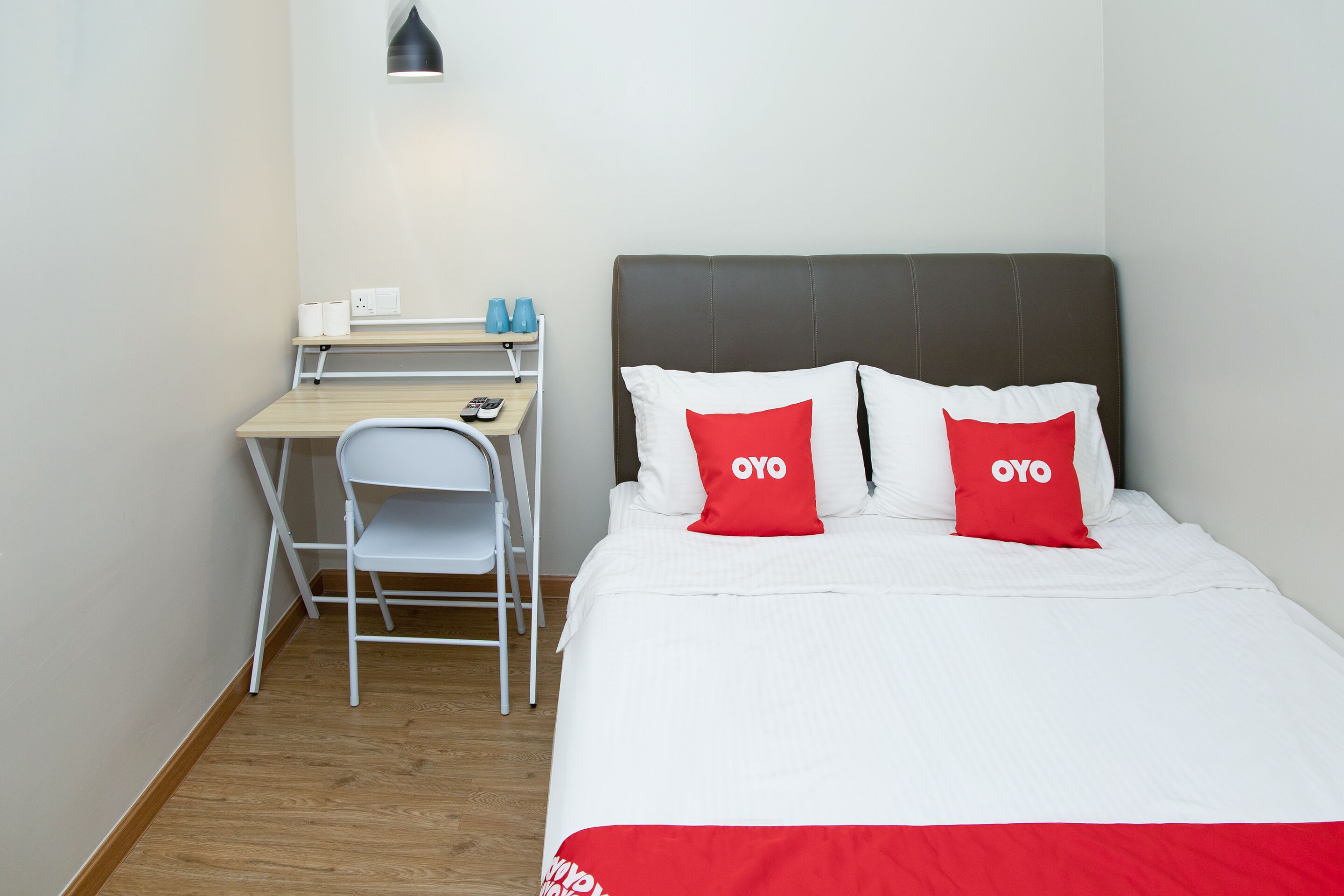 A hotel room with a bed, two red pillows with "OYO" text, a desk with a chair, a lamp, and a small shelf with toiletries.