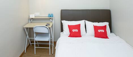 A hotel room with a bed, two red pillows with "OYO" text, a desk with a chair, a lamp, and a small shelf with toiletries.