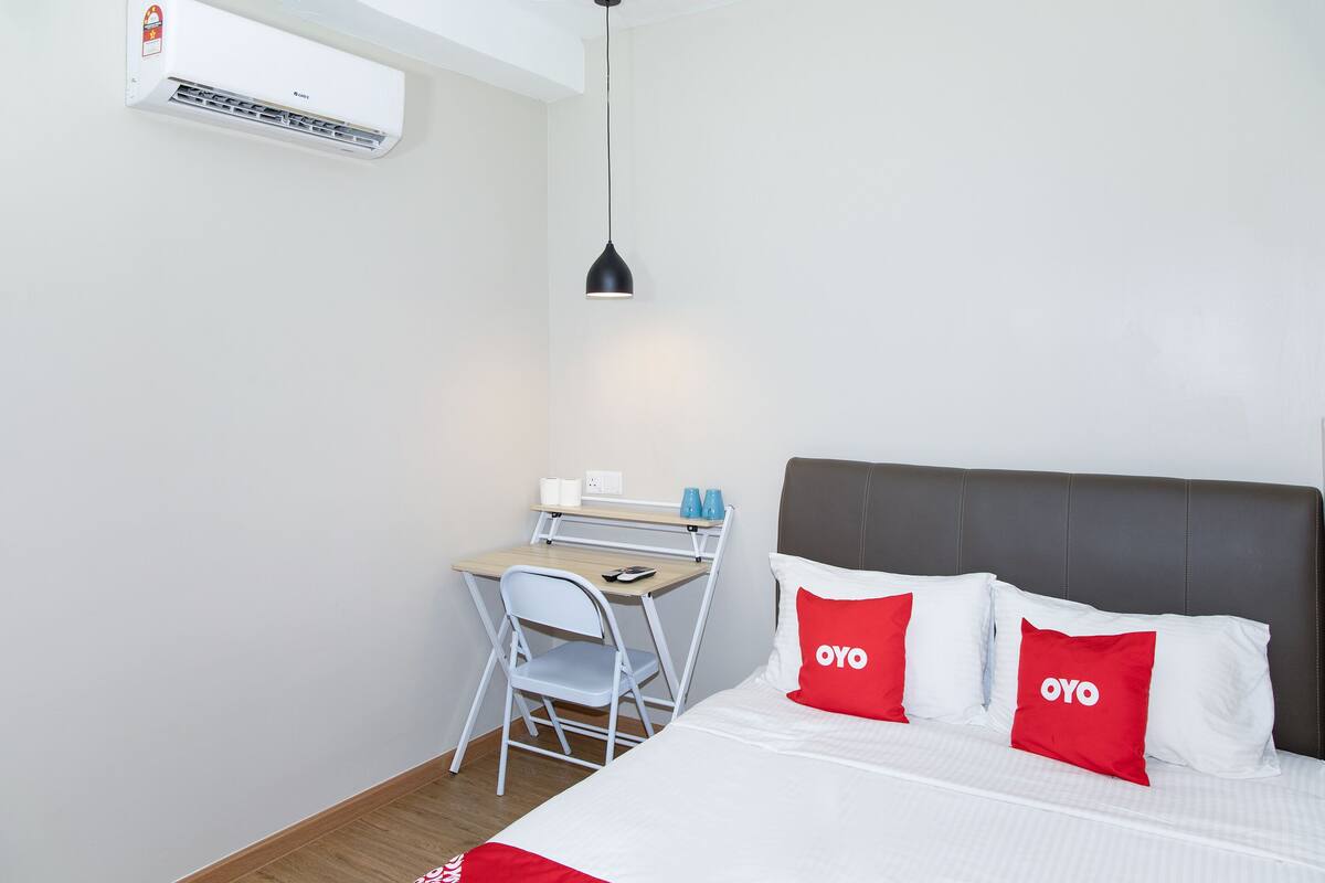 A compact hotel room with a bed, a desk, a chair, and a hanging light fixture.