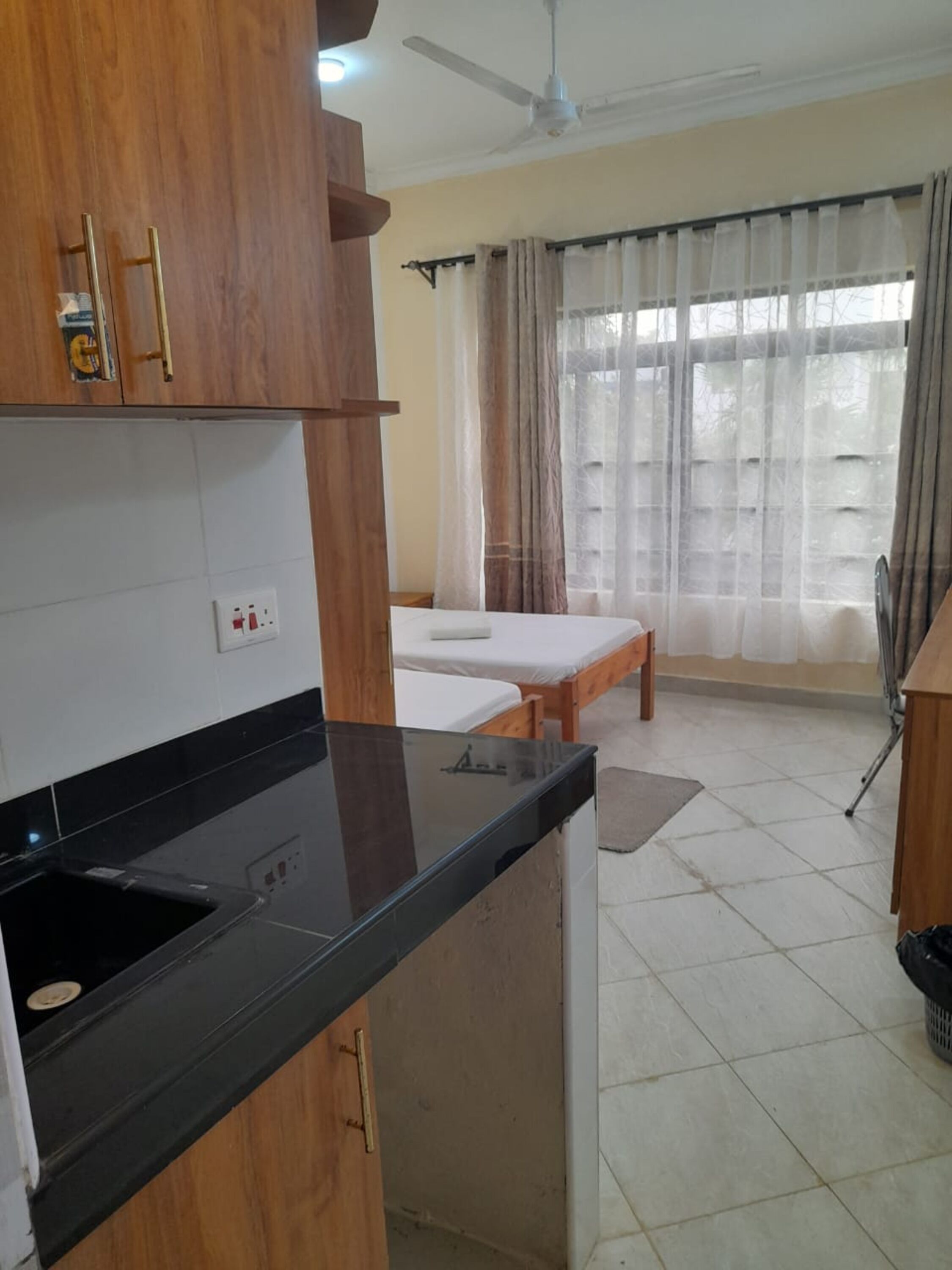 Una cocina compacta con encimera negra, armarios de madera y un lavamanos pequeño. En el fondo, se ve una cama con colchón blanco y cabecero de madera. Un ventilador de techo es visible sobre la cama.