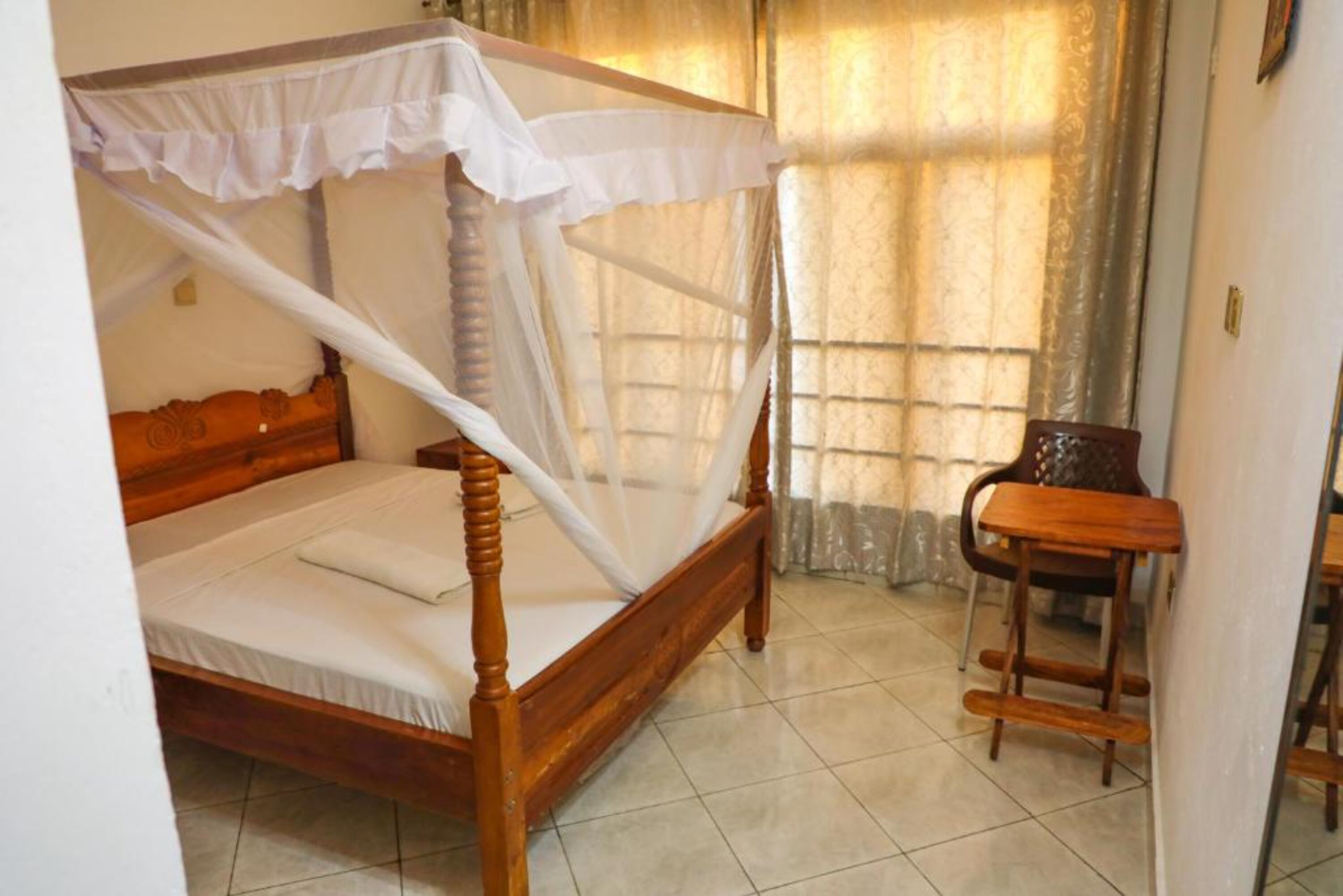 A single bed with a canopy, a wooden chair, and a tiled floor in a room with a window and curtains.
