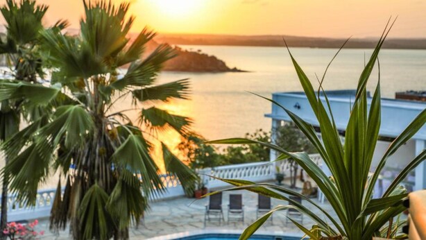 A poolside view with a sunset over the sea, palm trees, and a building with a white balcony. - Kilifi Blue House Resort (Kilifi)