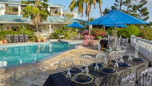 A poolside dining area with tables set for a meal, a pool, and a building with a blue roof and palm trees. - Kilifi Blue House Resort (Kilifi)