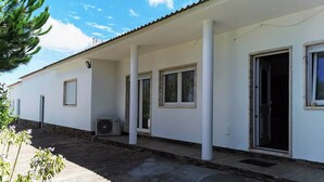 A single-story building with a flat roof, white exterior walls, and a covered porch area. There are multiple windows along the side of the building, and an air conditioning unit is visible on the porch. - Maison de Campagne au Calme (Ventosa)