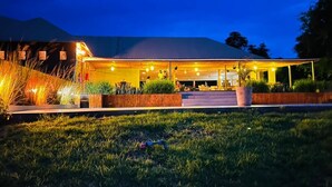 Un bâtiment moderne doté d’un espace extérieur couvert, qui pourrait être un restaurant ou un salon, avec une entrée bien éclairée et un parterre gazonné à l’avant.
